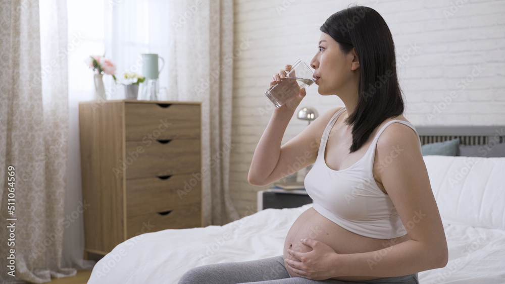 side portrait attractive asian expectant female wearing sportswear bra is drinking water while relaxing and gazing afar on bed in the bedroom. side portrait attractive asian expectant female wearing sportswear bra is drinking water while relaxing and gazing afar on bed in the bedroom.