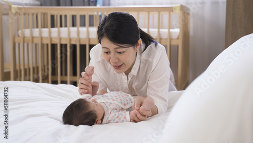 happy asian mom is enjoying time with her baby girl by holding and playing with the tiny feet and hands on the bed at background bright bedroom.