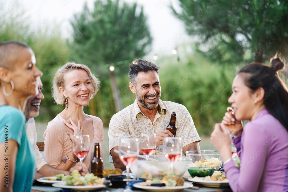 Group of friends having dinner around table in a garden on a summer evening, drinking wine, having fun talking and laughing Group of friends having dinner around table in a garden on a summer evening, drinking wine, having fun talking and laughing
