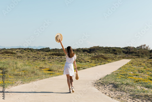 Back view of brunette woman holding sun hat and handbag while walking on beach.