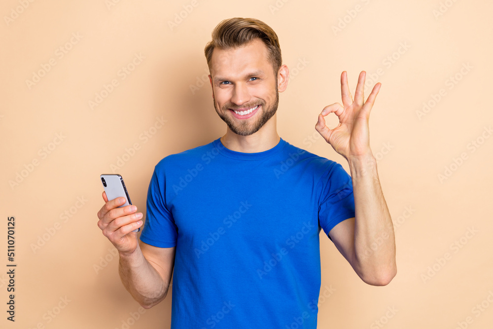 Photo of young guy use mobile show fingers okey symbol recommend promo perfect isolated over beige color background