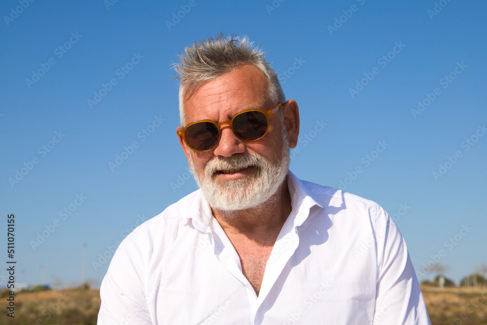 Handsome mature man with beard and grey hair is sitting pensively on a park bench while looking at the camera. The man is dressed in beige trousers, white shirt. Concept of old age and technology. Handsome mature man with beard and grey hair is sitting pensively on a park bench while looking at the camera. The man is dressed in beige trousers, white shirt. Concept of old age and technology.