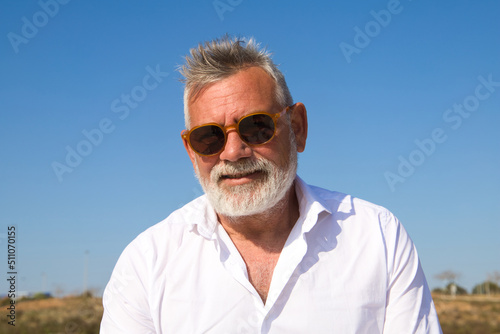 Handsome mature man with beard and grey hair is sitting pensively on a park bench while looking at the camera. The man is dressed in beige trousers, white shirt. Concept of old age and technology.