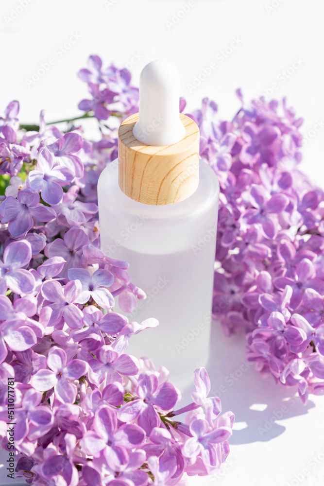 Skin care cosmetic product in glass bottle on white background with purple lilac flowers. Anti aging essence presentation. Natural ingredients for young healthy skin Skin care cosmetic product in glass bottle on white background with purple lilac flowers. Anti aging essence presentation. Natural ingredients for young healthy skin