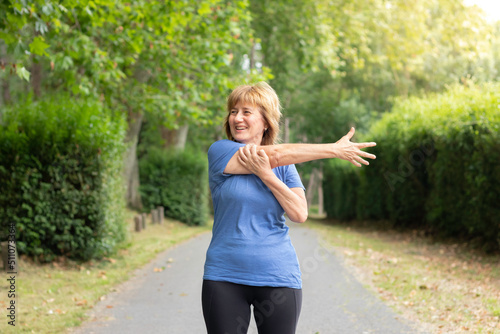 Mature woman in workout outfit smiling to the side streching her arm in the green park and trees around