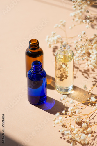 Open amber, blue and transparent bottles without cap with serum or essential oil with white flowers. Beige background with daylight and beautiful shadows. Beauty concept for face and body care