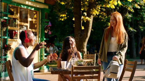 Two young women sitting at a table in the park are chilling as their two beautiful girl friends arrive and they hug to greet eachother