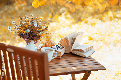 Bouquet of flowers, croissant, cup of tea or coffee, books on table in autumn garden. Rest in garden, reading books, breakfast, vacations in nature concept. Autumn time in garden on backyard photo