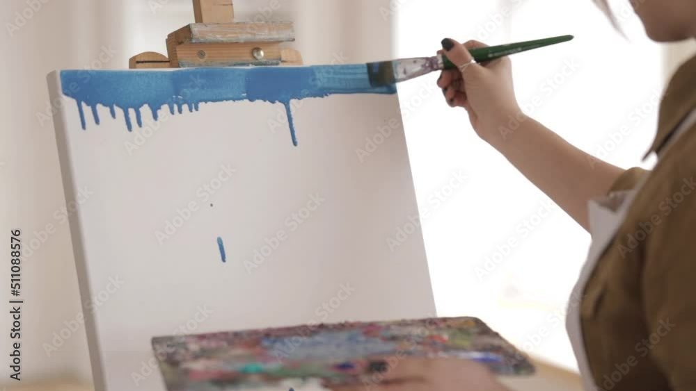 Woman is painting with brush on a canvas. Splitting the blue paint over