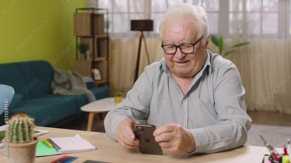 Capturing video of old man sitting on his working table at home in living room and looking through the smartphone and smiling Capturing video of old man sitting on his working table at home in living room and looking through the smartphone and smiling