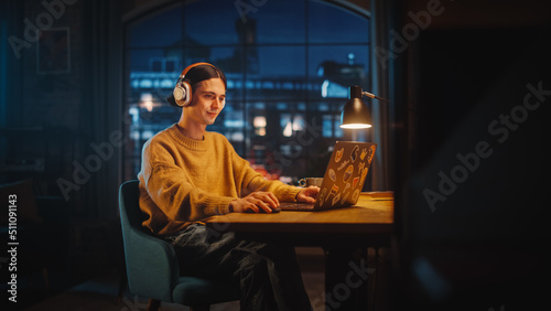 Young Handsome Man Opening Laptop Computer in Stylish Loft Apartment in the Evening. Creative Person Wearing Cozy Yellow Sweater and Putting On Headphones. Urban City View from Big Window.