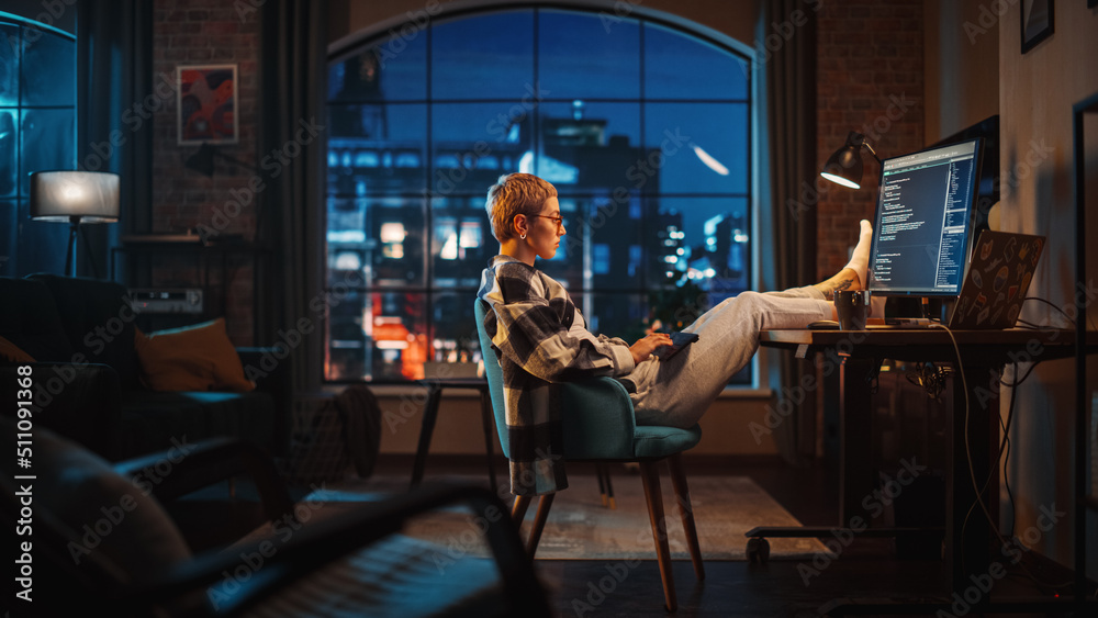 Young Woman Writing Code on Computer in Stylish Loft Apartment in the Evening. Creative Female Wearing Cozy Clothes, Resting Feet on Table and Using Wireless Keyboard. Urban City View from Big Window. Young Woman Writing Code on Computer in Stylish Loft Apartment in the Evening. Creative Female Wearing Cozy Clothes, Resting Feet on Table and Using Wireless Keyboard. Urban City View from Big Window.