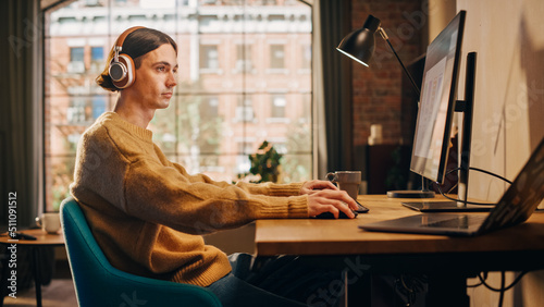 Young Handsome Man Sitting Down to Work on Desktop Computer in Sunny Stylish Loft Apartment. Creative Designer Wearing Cozy Yellow Sweater and Headphones. Urban City View from Big Window.