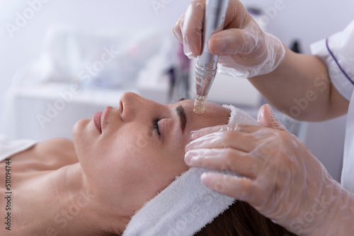 Fototapeta close up of Cosmetologist,beautician applying facial dermapen treatment on face of young woman customer in beauty salon