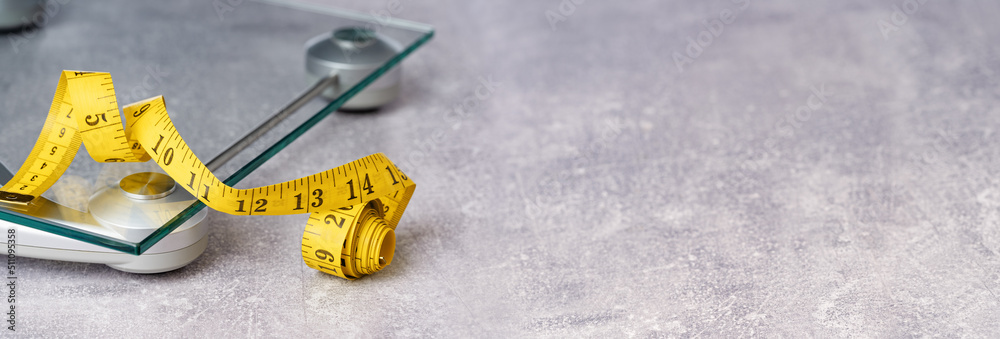 yellow tape measure on top of glass bathroom weighing scale on blue. Front view copy space. Healthy lifestyle, weight losing management and hydration concept banner