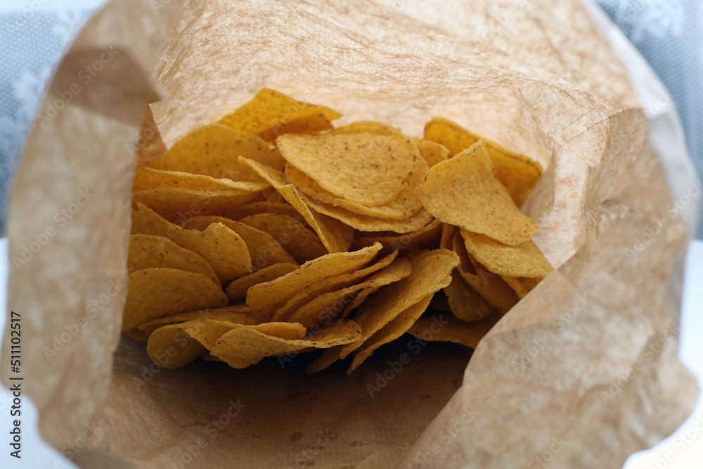 Brown paper bag full of yellow nachos on a white table. Mexican style appetizer. Unhealthy yet delicious food for snacking or eating. Crunchy vegetarian junk food. Closeup color image. Brown paper bag full of yellow nachos on a white table. Mexican style appetizer. Unhealthy yet delicious food for snacking or eating. Crunchy vegetarian junk food. Closeup color image.