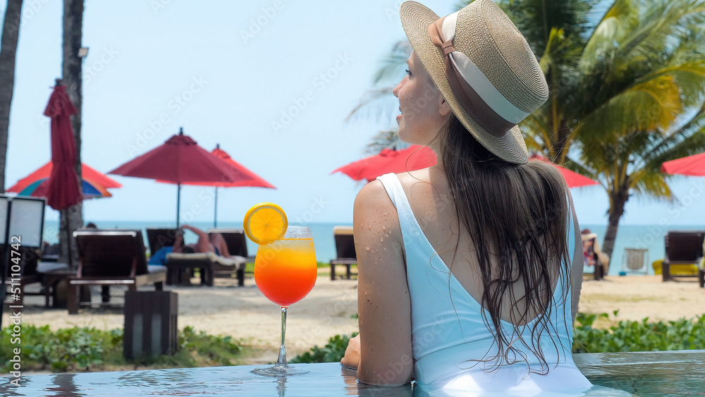 Fototapeta premium Woman in white swimwear and hat relax in swimming pool enjoy beautiful sea view with sun beds at sandy beach with tropical palms. Female traveler sunbathing in resort with glass of orange mocktail