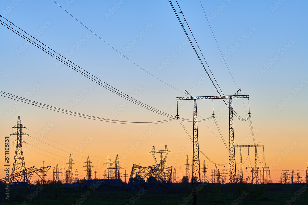 Dark silhouettes of power lines on fiery sunrise. High voltage electricity towers in field and fiery sunset. Concept of crisis of energy consumption, generation, and supply.