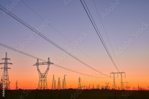 Dark silhouettes of power lines on fiery sunrise. High voltage electricity towers in field and fiery sunset. Concept of crisis of energy consumption, generation, and supply.