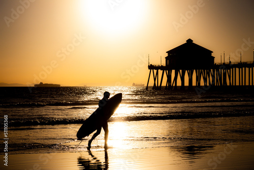 Surfer Walking on Beach Carrying Surfboard in Front of the Pier at Huntington Beach California at Sunset photo