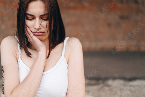 A Painful Looking Brunette Stands Against the Background of a Brick Wall. Girl Holds Her Hand to Her Cheek Squeezing Her Shoulders
