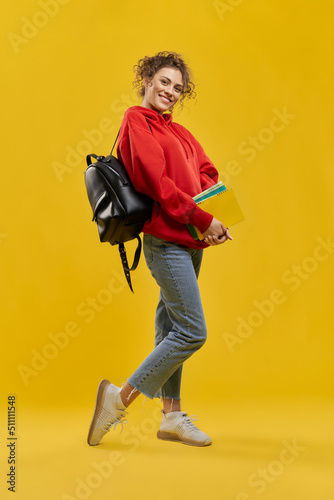 Pretty girl with backpack and notebooks, standing, shyly bent one leg on knee in studio. Front view of female student looking at camera, smiling, isolated on orange background. Concept of studying.