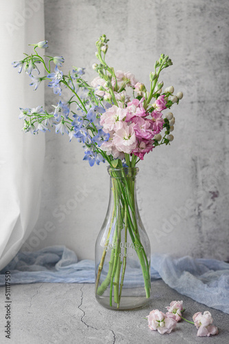 Fototapeta Naklejka Na Ścianę i Meble -  Bouquet of Blue Bellflowers, Pink Stock, and White Hypericum Berries in an Old Milk Bottle near a Window; Concrete Table and Background; Blue Fabric