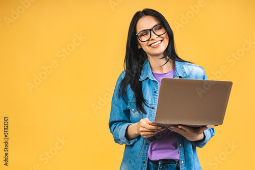 Portrait of happy young beautiful surprised woman with glasses standing with laptop isolated on yellow background. Space for text.