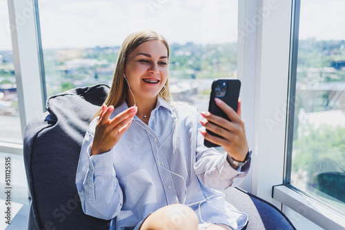 A young girl listens to music on headphones and watches something on a mobile phone. Modern woman lifestyle concept. Young woman sitting in coworking space, office interior