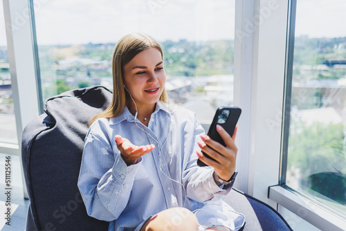 A young girl listens to music on headphones and watches something on a mobile phone. Modern woman lifestyle concept. Young woman sitting in coworking space, office interior