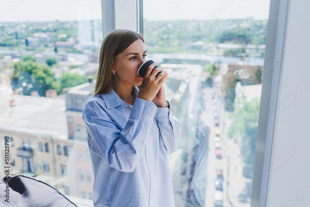 Fototapeta premium Happy woman drinks coffee and looks out the window in a cafe. A young smiling girl in glasses is standing by the window. Recreation, leisure and free time. The modern freelancer lifestyle