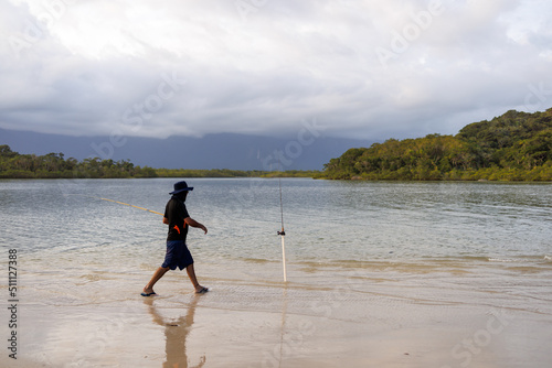 Homem andando em direção a sua vara de pesca fincada na margem do Rio  photo