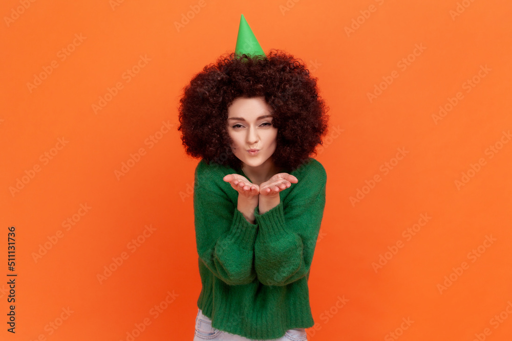 Happy birthday woman with Afro hairstyle wearing green casual style sweater thanks for the congratulations, sending air kisses to her guests. Indoor studio shot isolated on orange background. Happy birthday woman with Afro hairstyle wearing green casual style sweater thanks for the congratulations, sending air kisses to her guests. Indoor studio shot isolated on orange background.