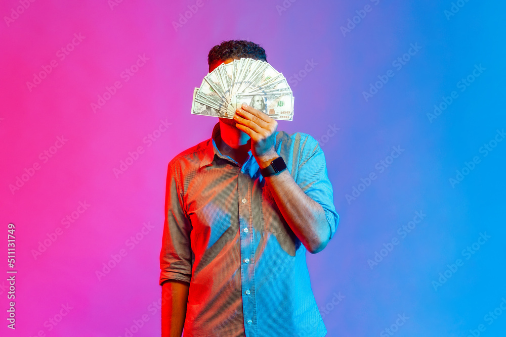 Portrait of man in shirt hiding face behind bunch of dollar banknotes, anonymous person holding money, lottery win, big profit. Indoor studio shot isolated on colorful neon light background.