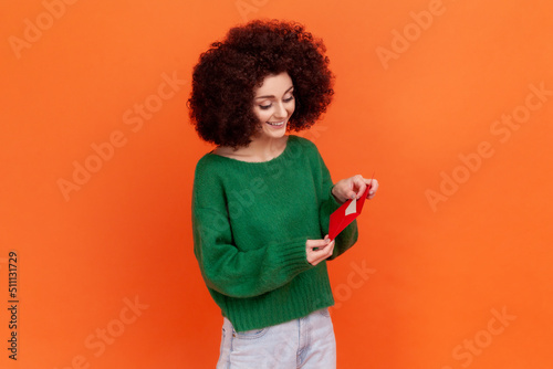 Portrait of positive female woman with Afro hairstyle wearing green casual style sweater standing holding red envelope, reading romantic letter. Indoor studio shot isolated on orange background.