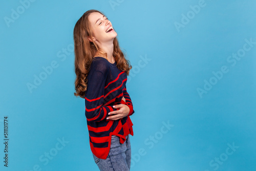 Hysterical laughter. Happy young woman wearing striped casual style sweater laughing out loud and holding belly, amused by humorous joke. Indoor studio shot isolated on blue background. photo