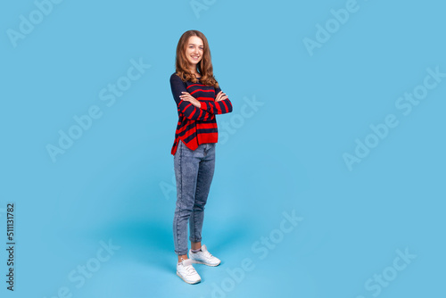 Full length portrait of good looking woman wearing striped casual style sweater and jeans standing with crossed arms, looking at camera. Indoor studio shot isolated on blue background.