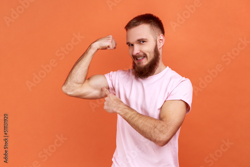 Portrait of proud strong bearded man pointing at raised hand demonstrating biceps, confident in body feeling powerful, wearing pink T-shirt. Indoor studio shot isolated on orange background.