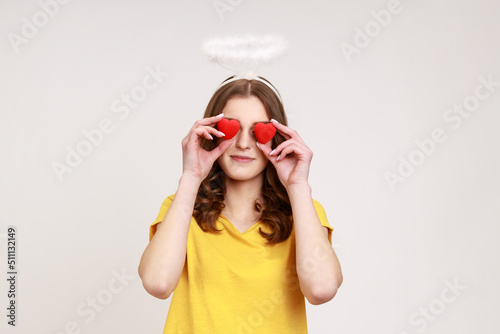 Portrait of funny angelic happy girl in yellow T-shirt with halo over head covering eyes with toy hearts and smiling, metaphor of look full of love. Indoor studio shot isolated on gray background.