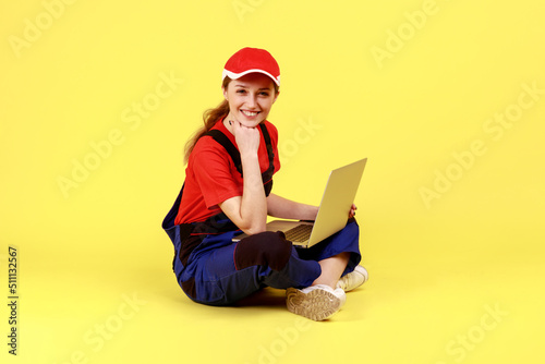 Foto Full length portrait of optimistic workwoman sitting on floor with crossed legs and working on notebook, looking at camera and holding chin