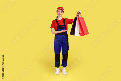Beautiful optimistic courier woman standing with shopping bags in hands, showing presenting parcels, expressing happiness, wearing overalls and cap. Indoor studio shot isolated on yellow background.