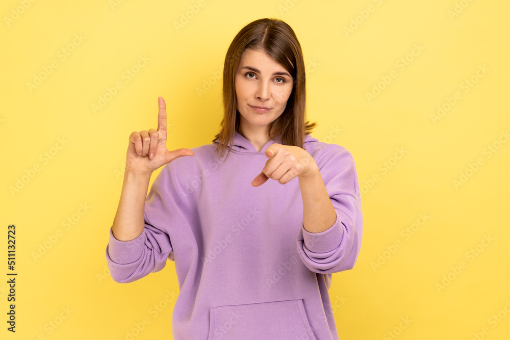 Portrait of frustrated young woman showing looser gesture and pointing at camera, looking with grumpy face, wearing purple hoodie. Indoor studio shot isolated on yellow background. Portrait of frustrated young woman showing looser gesture and pointing at camera, looking with grumpy face, wearing purple hoodie. Indoor studio shot isolated on yellow background.