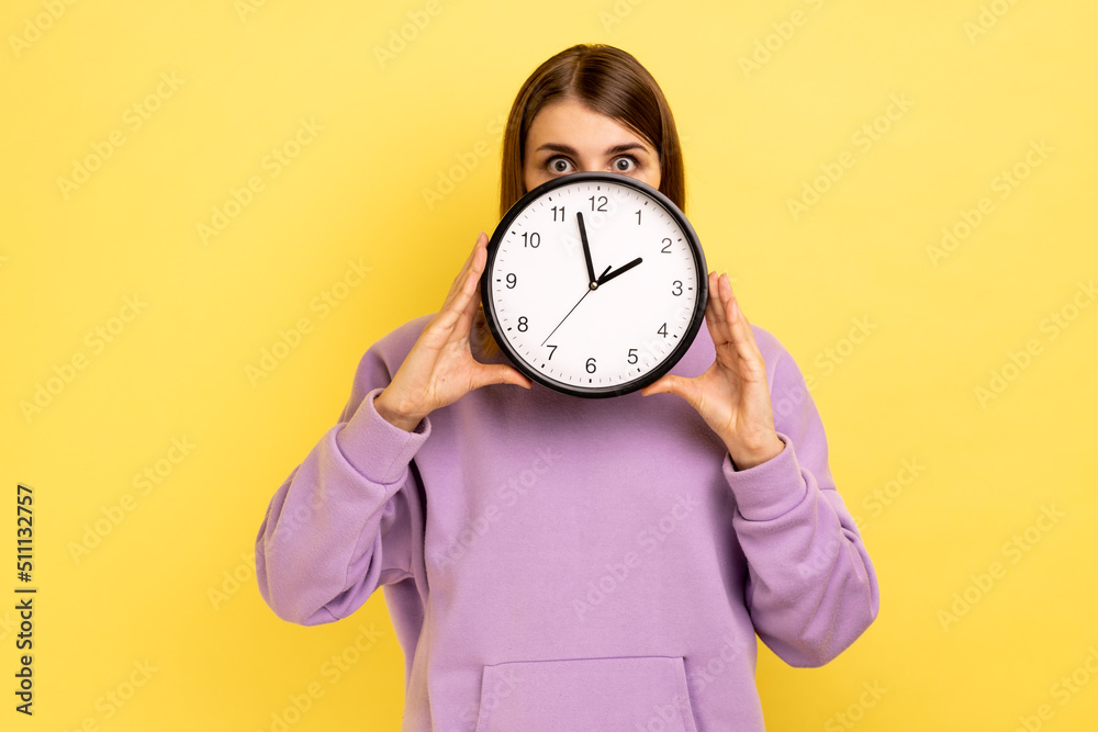 Astonished beautiful young adult woman covering half of face big wall clock, time management, schedule and meeting appointment, wearing purple hoodie. Indoor studio shot isolated on yellow background. Astonished beautiful young adult woman covering half of face big wall clock, time management, schedule and meeting appointment, wearing purple hoodie. Indoor studio shot isolated on yellow background.
