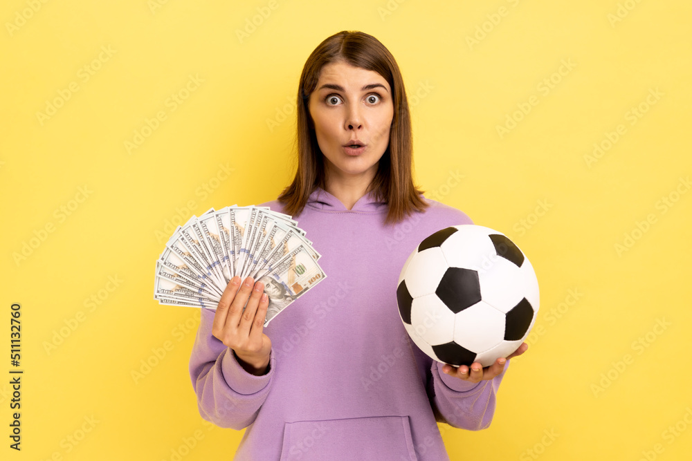 Portrait of woman with open mouth holding soccer ball and fan of dollars, amazed of football championship, betting, wearing purple hoodie. Indoor studio shot isolated on yellow background. Portrait of woman with open mouth holding soccer ball and fan of dollars, amazed of football championship, betting, wearing purple hoodie. Indoor studio shot isolated on yellow background.