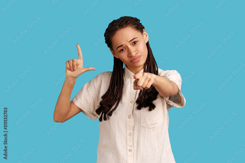 You are loser. Portrait of bossy serious woman with dreadlocks showing loser gesture and pointing at camera, accusing for failure, wearing white shirt. Indoor studio shot isolated on blue background. You are loser. Portrait of bossy serious woman with dreadlocks showing loser gesture and pointing at camera, accusing for failure, wearing white shirt. Indoor studio shot isolated on blue background.