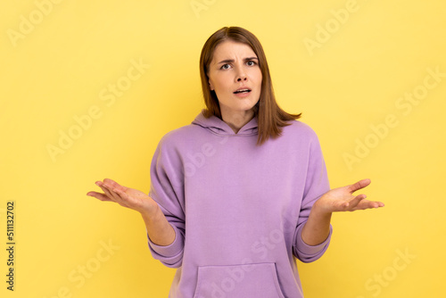 Portrait of unhappy stressed young adult woman standing raised arms and asking, expressing negative emotions, wearing purple hoodie. Indoor studio shot isolated on yellow background. photo