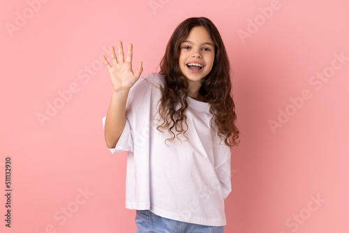 Portrait of friendly positive little girl wearing white T-shirt smiles toothily, raises palm greets friend, saying hello or goodbye. Indoor studio shot isolated on pink background.