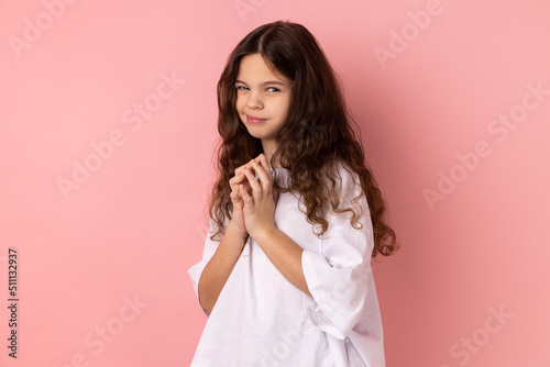 Portrait of cunning little girl in T- shirt having tricky plans, looking at camera with cheating face, smirking mysteriously, scheming cheats. Indoor studio shot isolated on pink background. photo