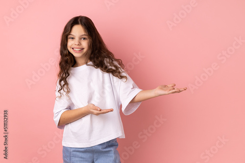 Obraz na plátně Portrait of kind-hearted generous little girl wearing white T-shirt showing welcome gesture and empty space on wall for your best advertising