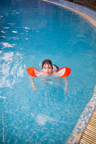 Boy in pool with orange swimming armlets smiling in blue water